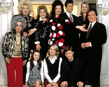 Cast of "The Nanny" posing in formal attire inside an elegant room, with ornate decorations in the background.