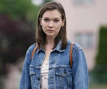 A young woman with shoulder-length hair wearing a jean jacket and a white floral top, standing outdoors with a neutral expression.