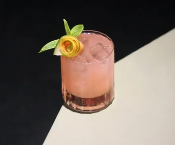 A pink cocktail in a glass with ice, garnished with a lemon peel rose and mint leaves on a black and cream surface.