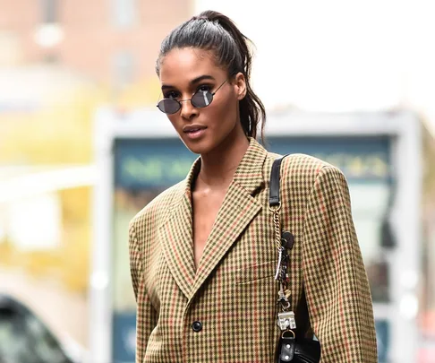 Model in a plaid blazer, sunglasses, and high ponytail on a city street.