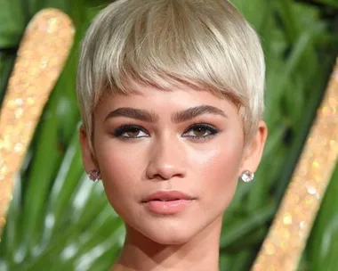 Woman with a blonde pixie cut hairstyle, wearing earrings, with a leafy background.