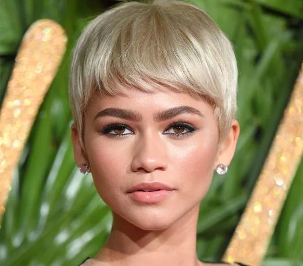 Woman with a blonde pixie cut hairstyle, wearing earrings, with a leafy background.