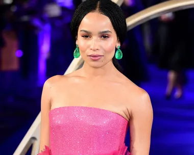 Zoe Kravitz wearing a sparkling pink strapless gown and green earrings at an event with a blue-lit background.