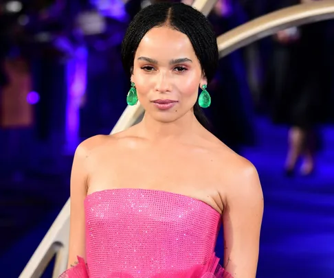 Zoe Kravitz wearing a sparkling pink strapless gown and green earrings at an event with a blue-lit background.