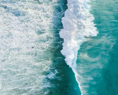 Aerial view of waves crashing on the ocean shore, displaying white foamy water and turquoise waves.