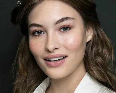 A young woman with long, wavy hair, light makeup, and clear skin smiles in a close-up portrait against a dark background.