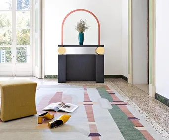 Room with modern decor, colorful geometric rug, yellow stool, vase on table, and yellow shoes and open book on the floor.