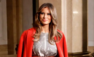 Woman with long light brown hair wearing a red coat, standing indoors with a neutral expression.