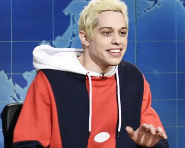 Pete Davidson on SNL's Weekend Update set, wearing a red, white, and blue hoodie, with a blonde hairstyle, smiling and speaking.