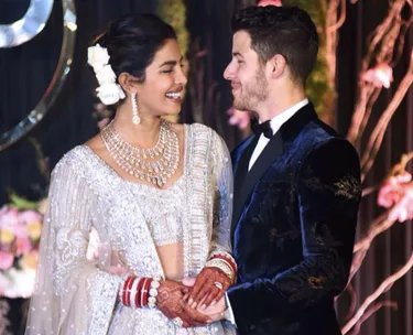 A bride and groom, dressed in traditional attire, smile at each other, holding hands at their wedding reception.