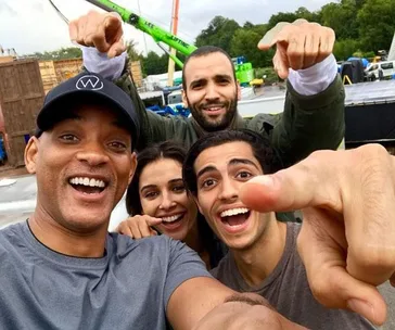 The cast of Disney's live-action Aladdin smiling and pointing at the camera on set.