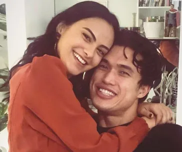 A smiling couple embraces in a cozy room with books and plants in the background.