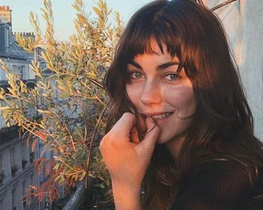 A woman with long hair and bangs smiles at the camera while standing on a balcony lined with plants in the evening light.