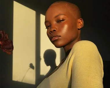 A person with a shaved head and a neutral expression in soft lighting with their shadow on the wall behind.