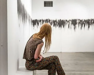 A woman with red hair, wearing patterned clothes, sits facing away in an art gallery with abstract black and white artwork on the wall.