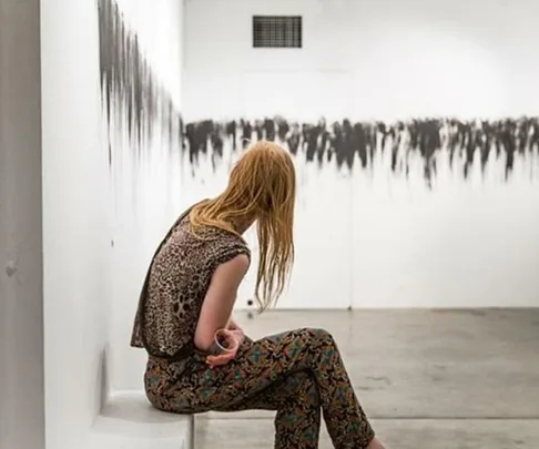 A woman with red hair, wearing patterned clothes, sits facing away in an art gallery with abstract black and white artwork on the wall.