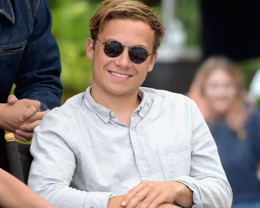 A man wearing sunglasses and a light button-up shirt smiles while sitting outdoors.