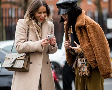 Two women in stylish coats, one holding a smartphone, both looking at the screen and smiling, with designer bags on their shoulders.