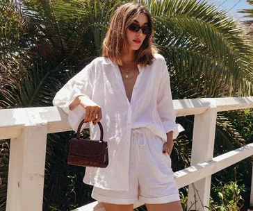 Woman in white linen shirt and shorts by Uniqlo, holding a small brown handbag, standing against a fence with palm trees in the background.