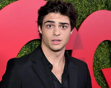 Noah Centineo, in a black suit, makes a surprised face at an event with a large "GQ" logo in the background.