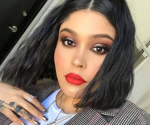 Woman with dark makeup and red lips, wearing a plaid jacket and silver rings, touches her hair while posing indoors.