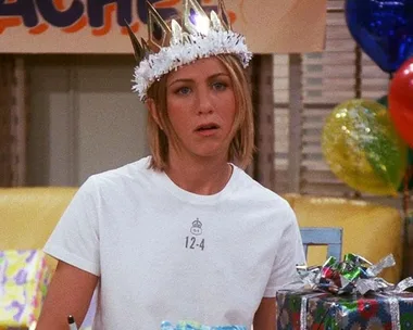 A woman wearing a crown and a t-shirt sits by gift-wrapped presents at a party, looking unamused.