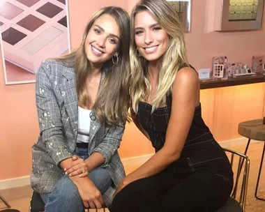 Renee Bargh interviewing Jessica Alba on a set with makeup products in the background.