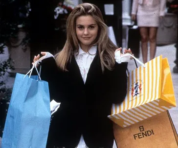 Woman holding shopping bags, dressed in a black blazer and white shirt, smiling outdoors.