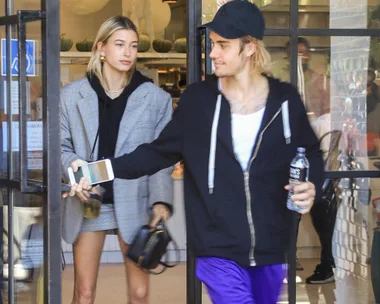 A couple exits a building; one holds a water bottle and a phone, the other wears a plaid blazer and holds a small bag.