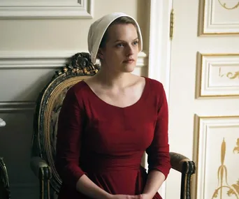 A woman in a red dress and white bonnet, seated in a room with ornate white walls and gold accents.