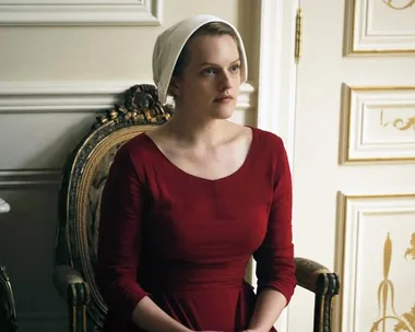 A woman in a red dress and white bonnet, seated in a room with ornate white walls and gold accents.