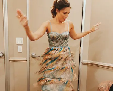 Woman in colorful fringed dress, arms raised, standing indoors near doors.