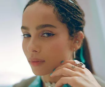 Person with braided hair and earrings, wearing a ring and light makeup, gazing softly at the camera.