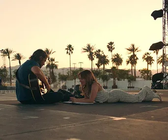 A man plays guitar while a woman writes on stage at sunset with palm trees in the background.