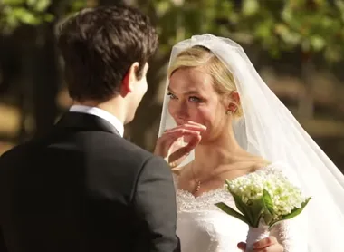 Bride in white gown holding bouquet, emotional, standing with groom in black suit outdoors.