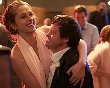 A man and woman happily dance together at a celebration, surrounded by smiling guests.