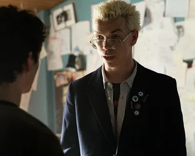 A man with bleached hair and glasses stands in front of another person in a room filled with papers on the walls.