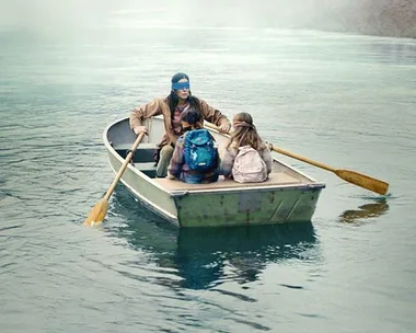 A blindfolded woman rows a boat with two children on a misty river scene from "Bird Box."