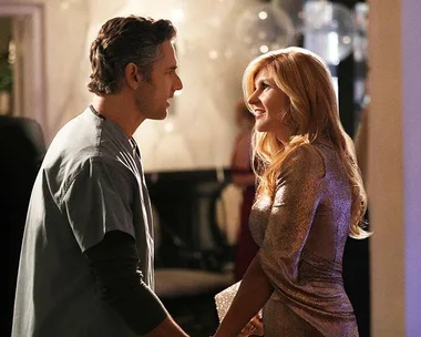 A man in scrubs and a woman in a sparkly dress smile at each other in a warmly lit room.