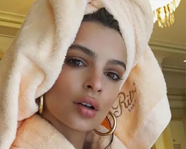 A person with a towel wrapped around their head and a bathrobe is looking towards the camera indoors.