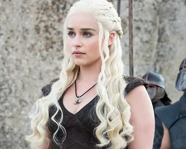 Daenerys Targaryen with long blonde hair, wearing a dark dress, standing outside in "Game of Thrones."