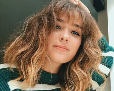 A woman with shoulder-length wavy hair, wearing a green striped sweater, looks at the camera with a slight smile.