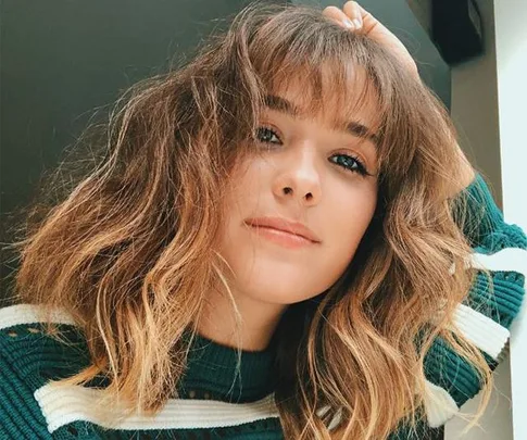 A woman with shoulder-length wavy hair, wearing a green striped sweater, looks at the camera with a slight smile.