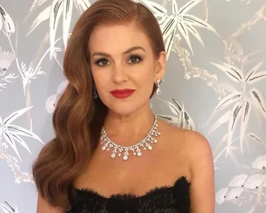 A woman in elegant attire with red hair and a statement necklace poses against a floral backdrop.