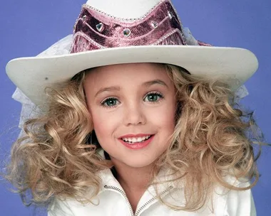 A young girl with curly blonde hair wearing a white cowboy hat and jacket, smiling against a blue background.