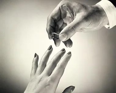 A man's hand places a wedding ring on a woman's finger against a neutral background.