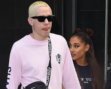 Man in pink shirt with sunglasses walks, while woman with long hair follows, both appearing relaxed.