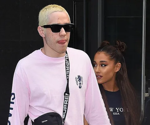 Man in pink shirt with sunglasses walks, while woman with long hair follows, both appearing relaxed.
