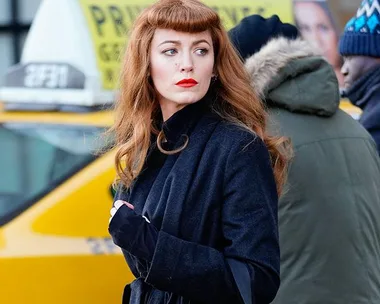 A woman with red hair and red lipstick stands in front of a yellow taxi, wearing a dark coat.