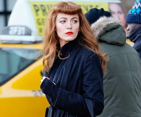 A woman with red hair and red lipstick stands in front of a yellow taxi, wearing a dark coat.